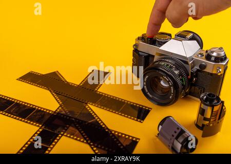 Ein Finger drückt den Auslöser einer antiken Kamera, umgeben von entwickelten 35-mm-Filmrollen. Die Szene wird durch einen gelben Hintergrund hervorgehoben. Stockfoto