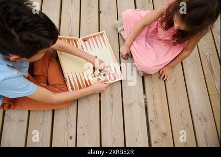 Zwei Kinder spielen ein Brettspiel auf einer Holzterrasse. Das Bild zeigt eine Draufsicht auf das laufende Spiel und hebt dessen Spaß und Konzentration hervor Stockfoto