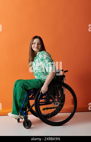 Eine selbstbewusste junge Frau in einem knallgrünen Outfit sitzt in einem Rollstuhl vor leuchtendem orangefarbenen Hintergrund und lächelt strahlend. Stockfoto