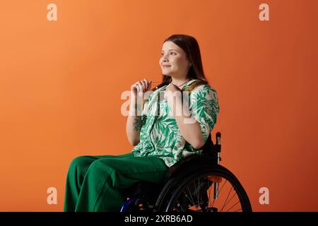 Eine junge Frau sitzt in einem Rollstuhl vor orangem Hintergrund, in lebendiger Kleidung gekleidet, die Selbstvertrauen und Freude ausstrahlt. Stockfoto