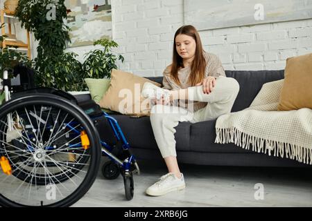 Eine junge Frau im Rollstuhl zieht ihre Turnschuhe an, während sie auf einer Couch in ihrem Wohnzimmer sitzt. Stockfoto