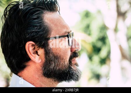 Seitliches Porträt eines kaukasischen erwachsenen Mannes mit Bart und Brille, der vor dem Hintergrund des Park-Natur-Geen mit defokussiertem Hintergrund schaut. Ernsthafte und professionelle männliche Leute im Freien Stockfoto