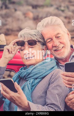 Fröhliches altes Seniorenpaar benutzt Handys im Freien zusammen mit rotem Auto im Hintergrund - Konzept von Reisereifen und moderner Technologie in älteren Rentnern glücklicher Lebensstil Stockfoto