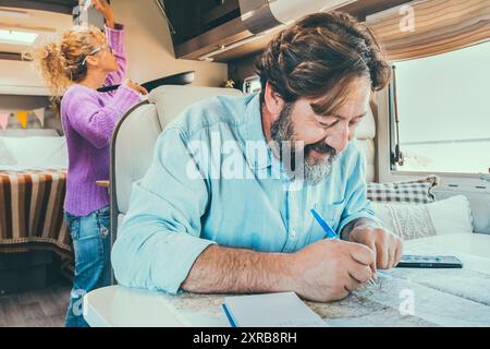 Bärtiger Mann, der den nächsten Reiseurlaub plant, sitzt am Tisch in seinem Wohnmobil. Frau im Hintergrund Kochen und Verwaltung der Lagerung. Urlaub im Urlaub Stockfoto