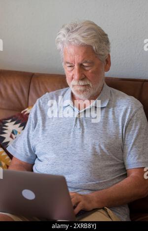 Ernster alter unzufriedener grauhaariger Mann sitzt auf dem Sofa und schaut auf den Bildschirm des Geräts, arbeitet am Laptop und sitzt allein zu Hause. Ein alter Geschäftsmann, der Nachrichten schreibt und geschäftliche Probleme über das Internet löst – reife Menschen und Verbindung Stockfoto
