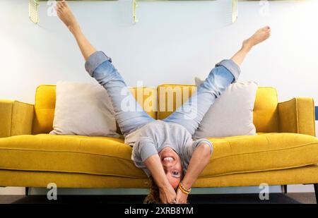 Fröhliche und fröhliche Erwachsene Frau legte sich in umgekehrter Position auf das Sofa, die Beine hoch und der Kopf runter, lächelte und hatte Spaß zu Hause - junge Dame mittleren Alters aktiver Lebensstil lachte viel auf der Couch Stockfoto