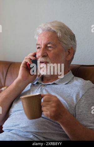 Ein alter älterer Mann zu Hause, der mit einem modernen Telefon spricht und Kaffee oder Tee trinkt, auf dem Sofa sitzt und die Technologie-Verbindung genießt - ein reifer Mann nutzt das Handy zu Hause, um sich zu entspannen und Spaß zu haben Stockfoto
