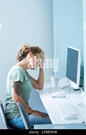 Gestresste und müde Erwachsene junge Frau am Desktop vor Computer und Online-Job Aktivität Tag Lebensstil berühren ihren Kopf für Kopfschmerzen Schmerzen - Büroarbeit - blaue Stimmung Stadt Farben Stockfoto