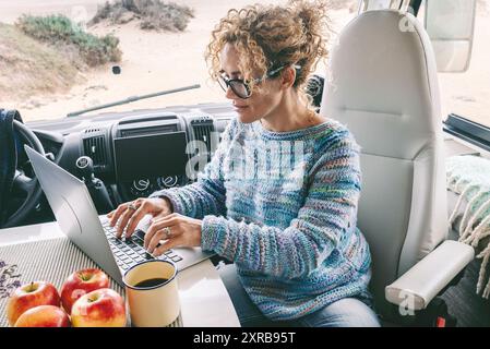 Leben und Arbeiten in Ihrem Wohnmobil auf Reisen und ein digitaler, nomadenfreier Lebensstil. Eine Frau, die in einem Wohnmobil sitzt, genießt es, sich zu entspannen und sich mit ihrem Laptop zu verbinden. Der Strand im Hintergrund vor dem Fenster Stockfoto