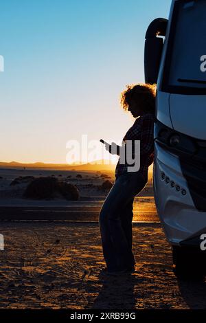 Frau in Silhouette gegen Sonnenuntergang schönes Licht. Reisen Sie mit einem Wohnmobil für einen Sommerurlaub oder einen freiheitlichen Lebensstil. Personen, die im Freien eine Telefonverbindung nutzen. Wohnmobil Vanlife Lifestyle Stockfoto