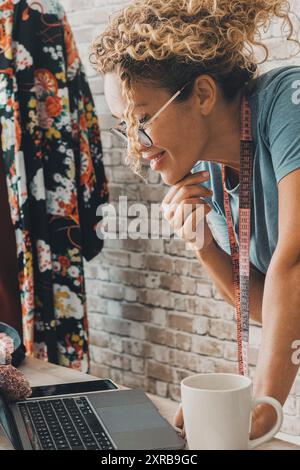 Moderne Stylistin Frau verwenden Computer und Verbindung für ihre Online-Arbeit. E-Commerce-Geschäft Unternehmer lächeln und schauen Laptop. Nähmaschine für handgemachte Kleidung Produktion. Schaffung von Künstlerjobs zu Hause Stockfoto