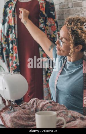 Frau streckt die Arme nach der Arbeit an der Nähmaschine zu Hause oder Design-Bekleidungsstudio. Weiblicher Stretching Körper nach Überlastung mit Kleidung Reparatur oder Produktion. Konzept der alternativen Arbeit in Innenräumen Stockfoto