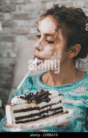 Junge, reife Frau, die zu Hause ein Mobiltelefon benutzt und einen Eiskuchen isst. Weiße Schönheitscreme-Behandlung im Gesicht. Indoor Freizeit weibliche Leute Aktivität.. Lady, die leckeres Essen isst und sich drinnen entspannt Stockfoto