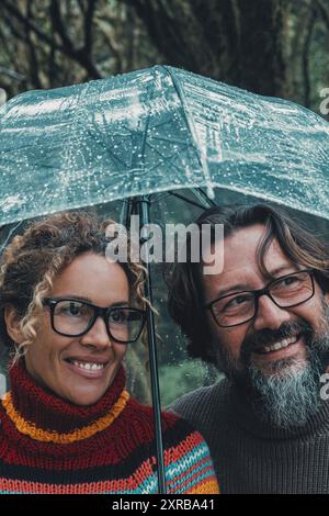 Beziehungs- und Liebespaar unter dem Regen lächelnd und genießen Outdoor-Aktivitäten an regnerischen Tagen geschützt unter einem transparenten Regenschirm. Mann und Frau umarmt und glücklich zusammen. Reife Menschen Stockfoto