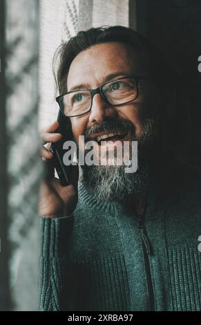 Ein glücklicher junger reifer Mann, der am Telefon spricht, lächelt und sich zu Hause am Fenster amüsiert. Konzept der Menschen- und Mobilfunk-Kommunikation. Männliche Person, die Handy benutzt, um mit Freunden zu kommunizieren Stockfoto