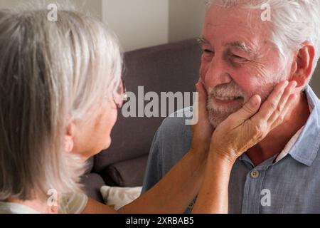 Eine alte Frau, die das Gesicht eines Reifen Mannes berührt, der zu Hause auf dem Sofa sitzt. Lebensstil im Alter. Reife Menschen genießen das Leben und die Beziehung. Dating. Pflege im Alter. Liebe Beziehung zusammen Stockfoto