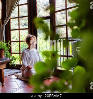 Lange, aufmerksame junge kaukasische Frau, die Mudra-Geste macht, in Lotusposition sitzend auf weichem Kissen auf dem Boden zu Hause sitzt. Friedliche Frau, die tief meditiert, allein Atemübungen macht. Stockfoto