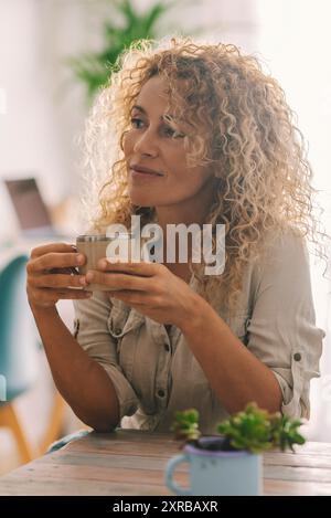 Eine attraktive Frau mit langen, blonden Locken genießt Zeit allein in der Freizeit zu Hause, trinkt Kaffee oder Tee und blickt weg, träumt und denkt nach. Ruhige weibliche Leute in entspannter Zeit drinnen Stockfoto