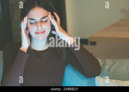 Eine Frau massiert ihren Kopf gegen starke Migräne, die auf dem Sofa sitzt, nachdem sie nachts bei Online-Aktivitäten einen Laptop benutzt hat. Menschen mit Kopfschmerzen und Stress wegen Misserfolg. Kopfschmerzkrankheit Stockfoto