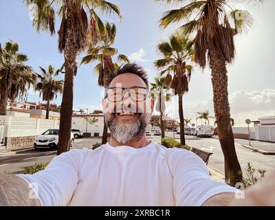 Ein reifer Mann macht Selfie bei Freizeitaktivitäten im Freien, spaziert auf der Straße an einem tropischen Ort mit Palmen im Hintergrund. Glückliche Touristen kommunizieren mit Handy Stockfoto