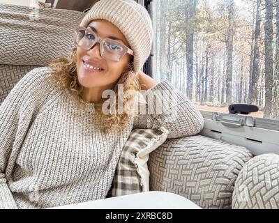 Schöne Frau mit Wollmütze schaut lächelnd in die Kamera. Die Dame sitzt im Wohnmobil und genießt ihren Urlaub im Wald Stockfoto