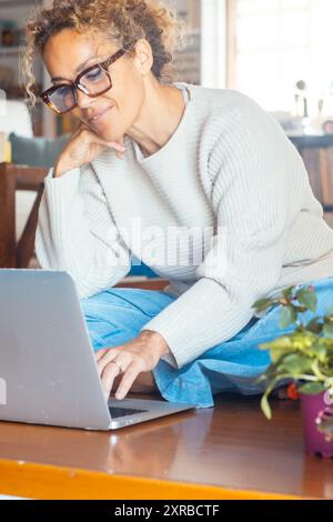 Eine Erwachsene Frau, die allein zu Hause am Laptop arbeitet und auf dem Holzboden sitzt. Drahtlose Verbindung und Technologie. Weibliche moderne Menschen, die Computer benutzen, um im Internet zu surfen und online zu einkaufen Stockfoto