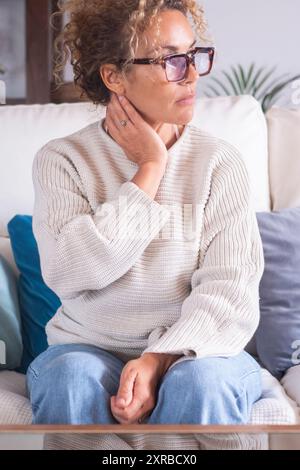 Eine Frau, die allein zu Hause auf dem Sofa sitzt, berührte und massierte ihren Hals schmerzhaft. Nachdenkliche weibliche Menschen und gesundes Konzept. Attraktive Gedanken Stockfoto