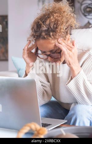 Ernsthafte, durchdachte, reife, freiberufliche Geschäftsfrau, die zu Hause am Laptop arbeitet, eine inländische Internetverbindung nutzt, wegblickt, denkt, entscheidet, sitzt auf dem Sofa am Computer, berührt das Kinn Stockfoto