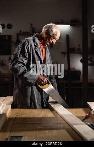 Ein alter Zimmermann sägt Holzbrett mit Metallsäge, ein pensionierter Mann, der in seiner Werkstatt Holzmöbel herstellt Stockfoto