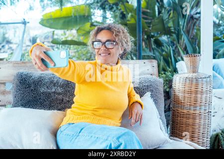 Eine Frau benutzt ihr Handy und entspannt sich allein im Garten auf einem Sofa. Porträt von fröhlicher weiblicher Schrift auf Handy. Frau macht Selfie. Reife junge Leute, die sich entspannen Stockfoto