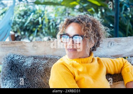 Glückliche blonde Frau mit Brille lächelt auf Sofa im Garten neben Haus Stockfoto