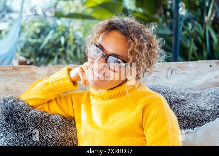 Glückliche blonde Frau mit Brille lächelt auf Sofa im Garten neben Haus Stockfoto