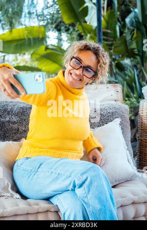 Eine Frau benutzt ihr Handy und entspannt sich allein im Garten auf einem Sofa. Porträt von fröhlicher weiblicher Schrift auf Handy. Frau macht Selfie. Reife junge Leute, die sich entspannen Stockfoto