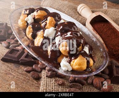 Schokoladen-Profiteroles mit Sahne, Kakaopulver, Kakaobohnen und Schokoladenstückchen. Stockfoto