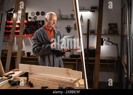 Zimmermann mit Laptop-Computer, der in seiner Tischlerei steht, mit verschwommenen Werkzeugen, die im Hintergrund an Bord hängen Stockfoto