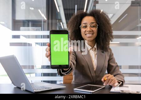Junge afroamerikanische Geschäftsfrau in professioneller Kleidung, die im Büro ein Smartphone mit grünem Bildschirm präsentiert. Selbstbewusste Frau lächelt und demonstriert Technologie mit Laptop und Dokumenten auf dem Schreibtisch Stockfoto