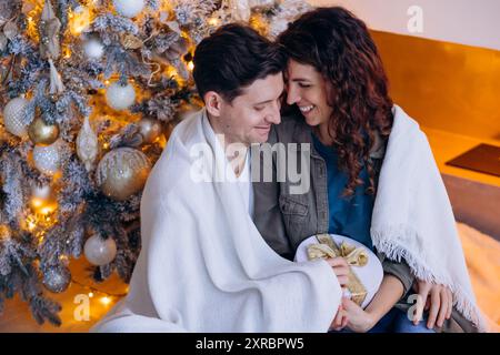 Glückliches Paar, das am Weihnachtsbaum sitzt, in Decke gehüllt, Geschenk hält, gemütliche Weihnachtsszene. Weihnachten feiern in einem gemütlichen Familienkreis, ein liebevoller Mann und eine Frau. Stockfoto