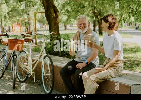 Seniorenpaar, das sich auf einer Holzbank ausruht und Wasser trinkt, nachdem es mit dem Fahrrad durch den Park gefahren ist Stockfoto