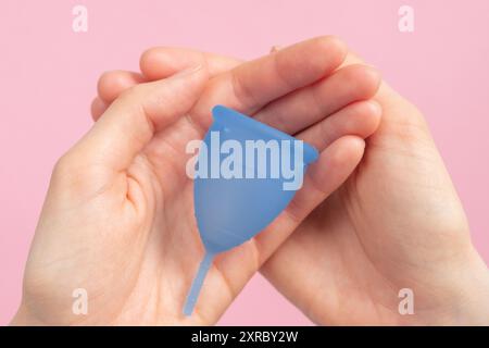 Frauenhände halten blaue Menstruationstasse vor rosa Hintergrund, Damenhygiene, nachhaltige Periodenpflege. Keine Verschwendung, schonend für die Umwelt. Stockfoto