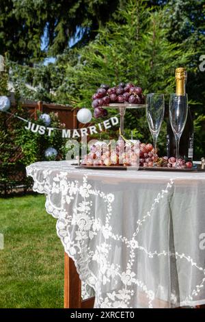 Ein Arrangement mit 2 Sektgläsern und einer Sektflasche neben roten Trauben auf einem silbernen Tablett steht auf einem Tisch mit einer weißen Tischdecke im Vordergrund, Vorbereitung auf eine Hochzeit im Garten, Schriftzug JUST Married im Hintergrund verschwimmt Stockfoto