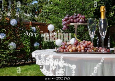 Ein Arrangement mit 2 Sektgläsern und einer Champagnerflasche neben roten Trauben auf einem silbernen Tablett im Vordergrund verschwimmt, Tisch mit weißer Tischdecke im Garten, Vorbereitung auf eine Hochzeit im Garten, Schrift GERADE verheiratet im Hintergrund scharf Stockfoto