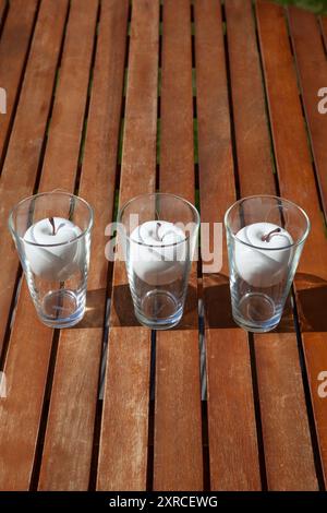 Weiße Plastikäpfel in Gläsern auf einem braunen Holztisch in der Sonne, Dekoration für eine Outdoor-Party Stockfoto