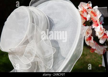 Ein weißer Damenhut mit Tüll hängt neben einem rot-weißen künstlichen Blumenkranz auf einem Holzstuhl im Garten zur Vorbereitung einer Hochzeit im Sommer Stockfoto