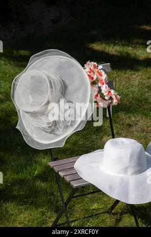 Ein weißer Damenhut mit Tüll hängt neben einem rot-weißen künstlichen Blumenkranz auf einem Holzstuhl im Garten, ein weißer Sommerhut liegt auf dem Sitz des Stuhls, Vorbereitung auf eine Sommerhochzeit Stockfoto