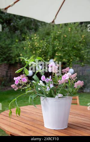 Ein weißer Cachepot mit frischen Blumen wie rosa Sackblumen, weißen und rosa Wicken und grünen Blättern steht als Blumenarrangement auf einem Holztisch im Garten, ein Apfelbaum mit hellgrünen Äpfeln steht verschwommen im Hintergrund Stockfoto