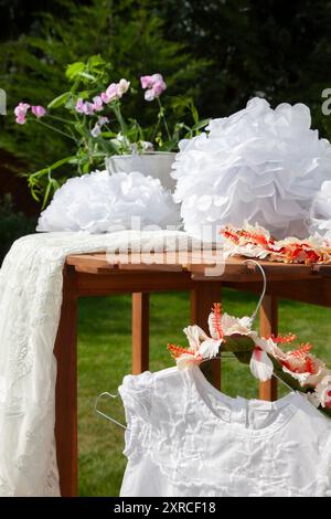 Nahaufnahme eines weißen Kinderkleides mit Spitze und einem Blumenkranz als Kopfschmuck, der an einem Kleiderbügel am Tisch hängt, Vorbereitungen für eine Hochzeit, Hochzeitsvorbereitung im Freien Stockfoto