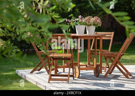 Gartenmöbel auf einer Holzterrasse im letzten Sonnenlicht des Tages, Sitze in der Abendsonne im grünen Garten im Spätsommer ragen die grünen Blätter einer Weide im Vordergrund ins Bild Stockfoto