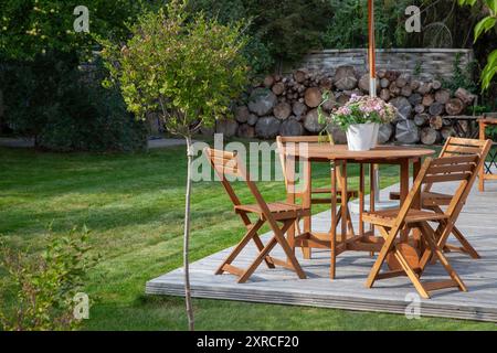 Gartenmöbel auf einer Holzterrasse im letzten Sonnenlicht des Tages, Sitzen in der Abendsonne im grünen Garten im Spätsommer, ein kleiner Fliederbaum im Vordergrund Stockfoto