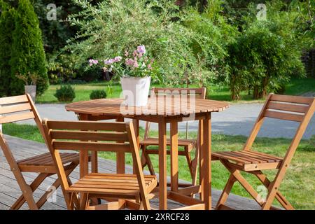 Close-up Holztisch mit Gartenstühlen auf einer Außenterrasse, Gartenmöbel auf einer Holzterrasse, Blumenarrangements auf dem Holztisch, grüner Garten im Spätsommer, Urlaub zu Hause, verschwommener Hintergrund mit grünen Büschen und Bäumen Stockfoto