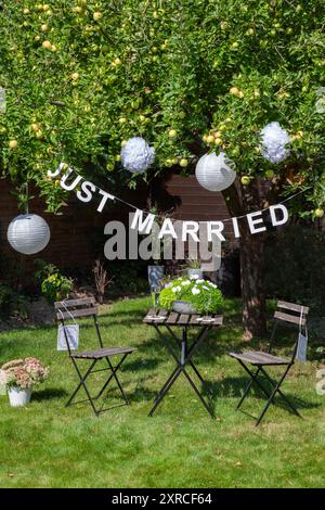 Eine Girlande mit weisen Buchstaben und die Inschrift "gerade verheiratet" hängt an einem Apfelbaum mit grünen Äpfeln im Garten, Schilder hängen von den beiden Stühlen. Stockfoto